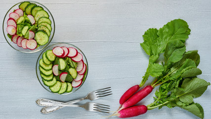 Summer detox salad with radish, cucumber, spinach in two glass bowls on the gray kitchen table. Healthy diet breakfast decorated with bunch of organic radishes and silver forks. Top view