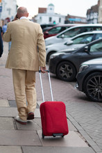 Male Tourist In Cream Creased Linen Suit White Shoes And Bald Head Walks Along Sidewalk Pavement In Typical English Town Talking On Mobile Phone And Pulling Bright Red Flight Bag With Handle
