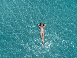 Fototapeta  - Beautiful young woman floating in crystal clear water, aerial shot
