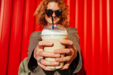 Hipster young woman with curly red hair holding coffee against red wall. Close-up of a drink in plastic cup