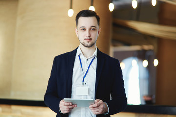 Waist-up portrait of business conference attendee looking at camera with tablet computer in his hands, empty badge around his neck