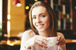 Content young woman holding cup of coffee and smiling at camera while relaxing in cafe.