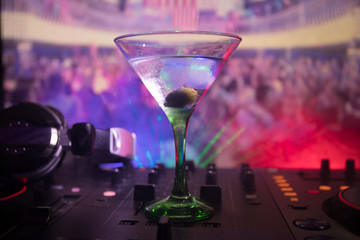 Glass with martini with olive inside on dj controller in night club. Dj Console with club drink at music party in nightclub with disco lights. Close up view