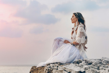 beautiful young stylish boho woman sitting on the beach at sunset