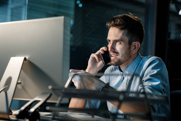 Wall Mural - Bearded handsome man speaking on smartphone while watching computer in office late at night. 