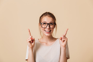 Poster - Image of blonde woman 20s with tied hair wearing basic t-shirt and eyeglasses smiling while pointing fingers upward, isolated over beige background in studio