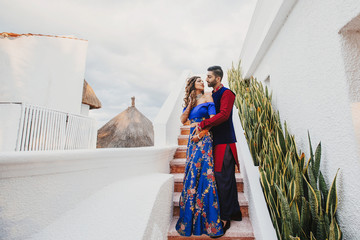 Wall Mural - Gorgeous Hindu groom in blue and red sherwani and bride in blue lehenga pose on the stairs in white house with greenery