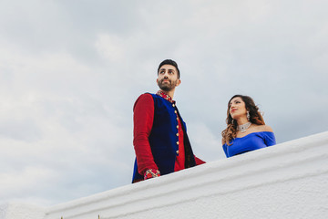 Gorgeous Hindu groom in blue and red sherwani and bride in blue lehenga pose on the stairs in white house with greenery