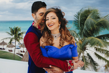 Wind blows around Hindu groom in blue sherwani and bride in lehenga posing in white house with gorgeous seaside view behind them