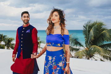 Wall Mural - Wind blows around Hindu groom in blue sherwani and bride in lehenga posing in white house with gorgeous seaside view behind them