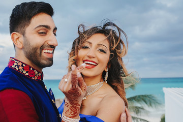 Wind blows around Hindu groom in blue sherwani and bride in lehenga posing in white house with gorgeous seaside view behind them