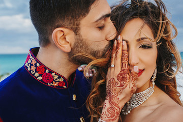 Wind blows around Hindu groom in blue sherwani and bride in lehenga posing in white house with gorgeous seaside view behind them