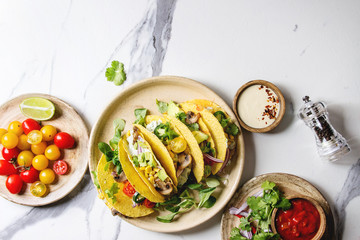 Wall Mural - Variety of vegetarian corn tacos with vegetables, green salad, chili pepper served on ceramic plate with tomato and cream sauces with ingredients above over white marble background. Top view, space.