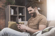 © Yakobchuk Olena - Joyful news. Side view of cheerful young bristled man is sitting on comfortable couch at home and reading funny message on his modern mobile phone. He is looking at screen with smile