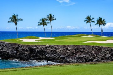 Sunny day on a tropical golf course fairway with the putting green in the distance surrounded by palm trees and sand traps, lava rock, blue pacific ocean, and blue sky and white clouds in background