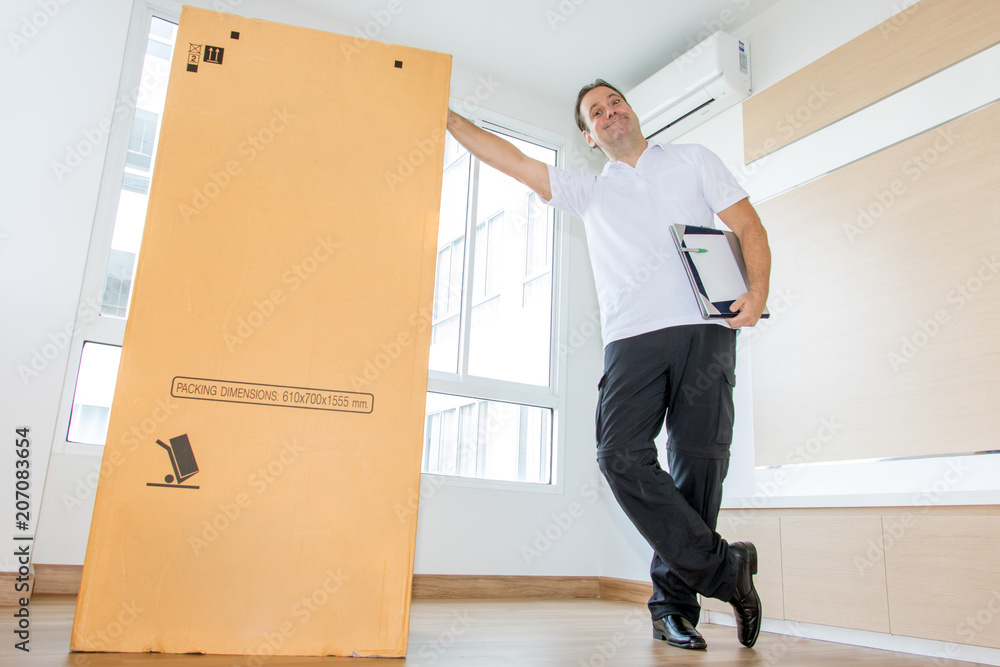 A man standing beside a large package in an empty room. The postman delivers the parcel to the new apartment. - obrazy, fototapety, plakaty 