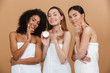 Beauty photo of three smiling multiracial women with different types of skin: caucasian, african american and asian girls, applying face cream together isolated over beige background