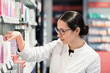 © Kzenon - Side view close-up portrait of a dedicated female pharmacist standing in front of the pharmacy shelves with various products, while thinking to make the best choice