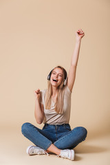 Poster - Happy young girl 20s rejoicing and raising up arm while sitting on floor with legs crossed and listening to music via wireless headphones, isolated over beige background