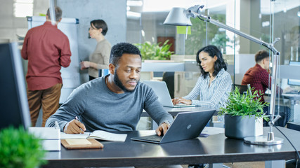 Busy International Office, African-American Man Working at His Desk on a Laptop, He Writes in Reminder His Notebook. In Background Creative Young People Working.