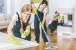Young member of a cleaning crew wearing green overalls and yellow gloves wiping a white table in apartment interior with the rest of the team in blurred background