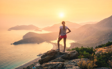 Wall Mural - Young woman standing on the top of rock and looking at the seashore and mountains at colorful sunset in summer. Landscape with girl, sea, mountain ridges and orange sunlight. Travel. Oludeniz, Turkey