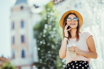 Beautiful young woman in sunglasses and hat in cafe drinking mojito refreshing cocktail while talking on the phone in the street on sunny day