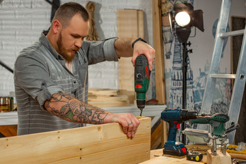 profession, carpentry, woodwork and people concept - carpenter with electric drill drilling wood plank at workshop