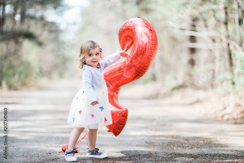 Blonde white female toddler wearing a grey and white dress with multi colored butterflies pointing at a red number two balloon smiling at camera Poster