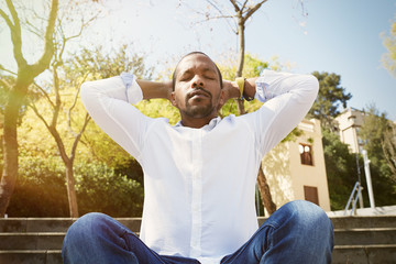 Young american african businessman in white shirt put his hands on his head and taking a deep breath outside corporate office.Business yoga and stress free environment.Peace of mind concept