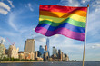 © lazyllama - Colorful rainbow gay pride flag fluttering in the breeze against a sunny city skyline