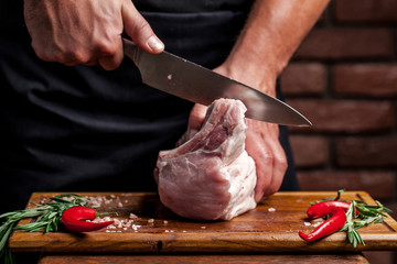 The concept of cooking meat. The man chef, the butcher, cuts raw meat with beef, lamb, veal, holding a knife in his hand, on a wooden table, next to lie raw vegetables, cherry, parsley, dill.