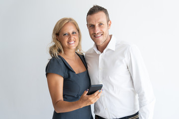 Wall Mural - Portrait of smiling mid adult husband and wife standing together, looking at camera and smiling, Woman holding mobile phone. Mobile technology concept