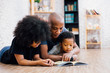 © twinsterphoto - African American father reading a fairy tale fable story for kids at home. Happy family lying on the floor indoors