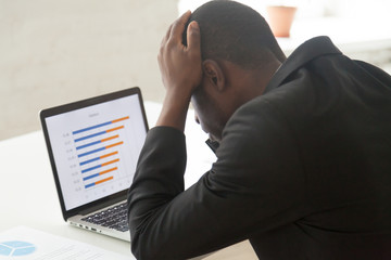 Wall Mural - Disappointed stressed African American manager holding head in hands looking at laptop screen, observing falling rates online, witnessing business collapse, company failure. Bankruptcy, loss concept