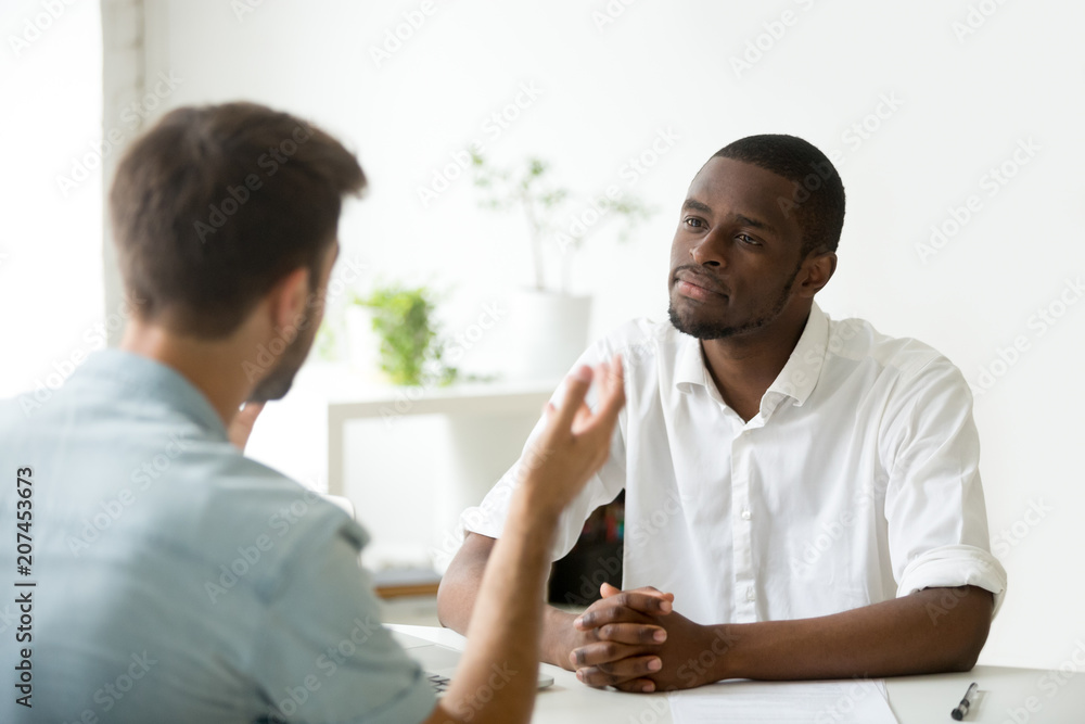 African American employer listening attentively to caucasian job applicant talking at work interview, being friendly and interested to candidate. Concept of recruiting, employment, hiring - obrazy, fototapety, plakaty 