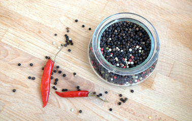 Still life with red paprika chilli pepper and black pepper seeds inside round glass jar and scattered on wooden table background top view closeup