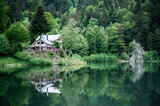 Fototapeta  - A house with red shutters. Around the house is a green forest. In front of the house is a lake.