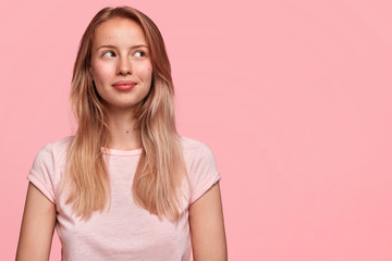 Wall Mural - Beautiful cute young female with pensive look, concentrated upwards, has dreamy positive expression, thinks about something pleasant and unforgettable, poses against pink background. Copy space