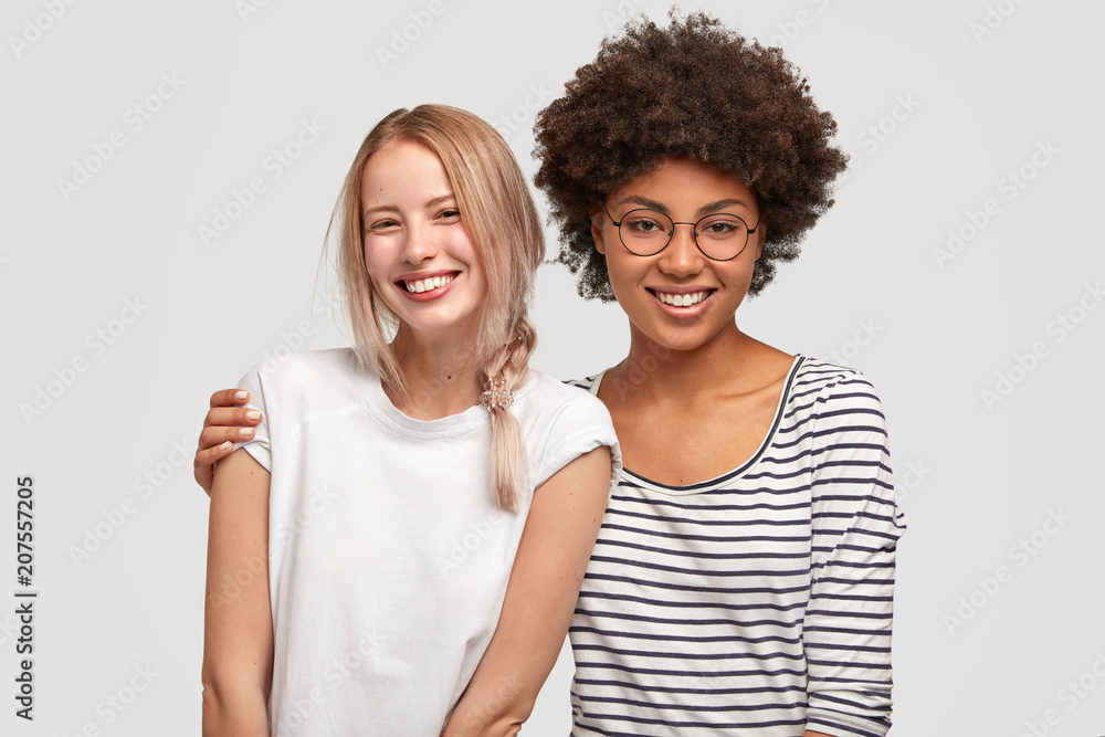 Two beautiful mutiethnic delighted women have good relationship and truthful friendship, embarce each other and smile gladfully, happy to meet after long time. Interracial friendship concept - obrazy, fototapety, plakaty 