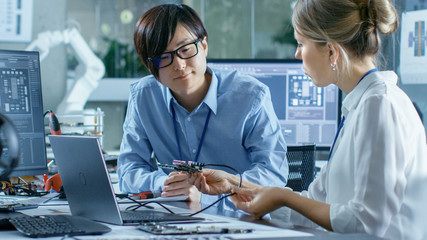 Wall Mural - Two Computer Engineers Talking, They Connect Printed Circuit Board to a Laptop and Start Coding. In the Background Computer Science Research Laboratory with Robotic Arm Model.