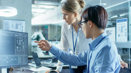 Asian Scientist Sitting at His Desk Consults Senior Engineer about Sophisticated Coding and Programming. In the Background Computer Science Research Laboratory with Robotic Arm Model.