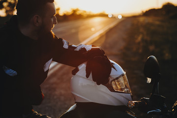 Wall Mural - Side view of a confident caucasian traveler sitting on his bike while resting near the road and looking at the sunset with a hand on his helmet while traveling.