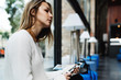 © JKstock - Portrait of charming blonde woman sitting beside a big window of a modern coffee shop with a mobile phone in her hands. Thoughtful student girl holding smartphone while sitting on a blurred background