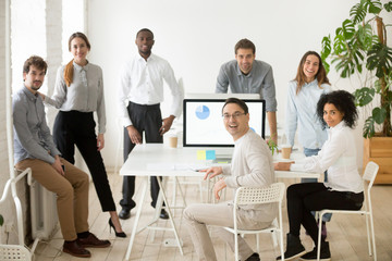 Poster - Young casual multiracial team and middle aged leader or coach looking at camera, smiling, standing near office desks in coworking space, posing for company business portrait. Team spirit, cooperation