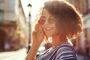 Outdoor close up portrait of young beautiful stylish happy smiling curly girl wearing red narrow oval sunglasses, posing in street. Sunny day light. Summer fashion concept. Copy, empty space for text