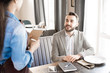 © Seventyfour - Cheerful handsome bearded young businessman in casual outfit smiling at waitress while making order and sitting at table with diary and smartphone in restaurant