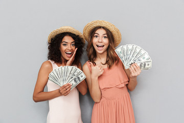 Canvas Print - Portrait of two successful multiethnic women, caucasian and african american girls wearing straw hats holding fans of money 100 dollar banknotes, isolated over gray background