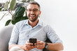 © Drobot Dean - Image of smiling employer guy in white shirt sitting in office armchair near green plant, and using smartphone for work