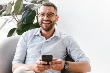 Image of smiling employer guy in white shirt sitting in office armchair near green plant, and using smartphone for work